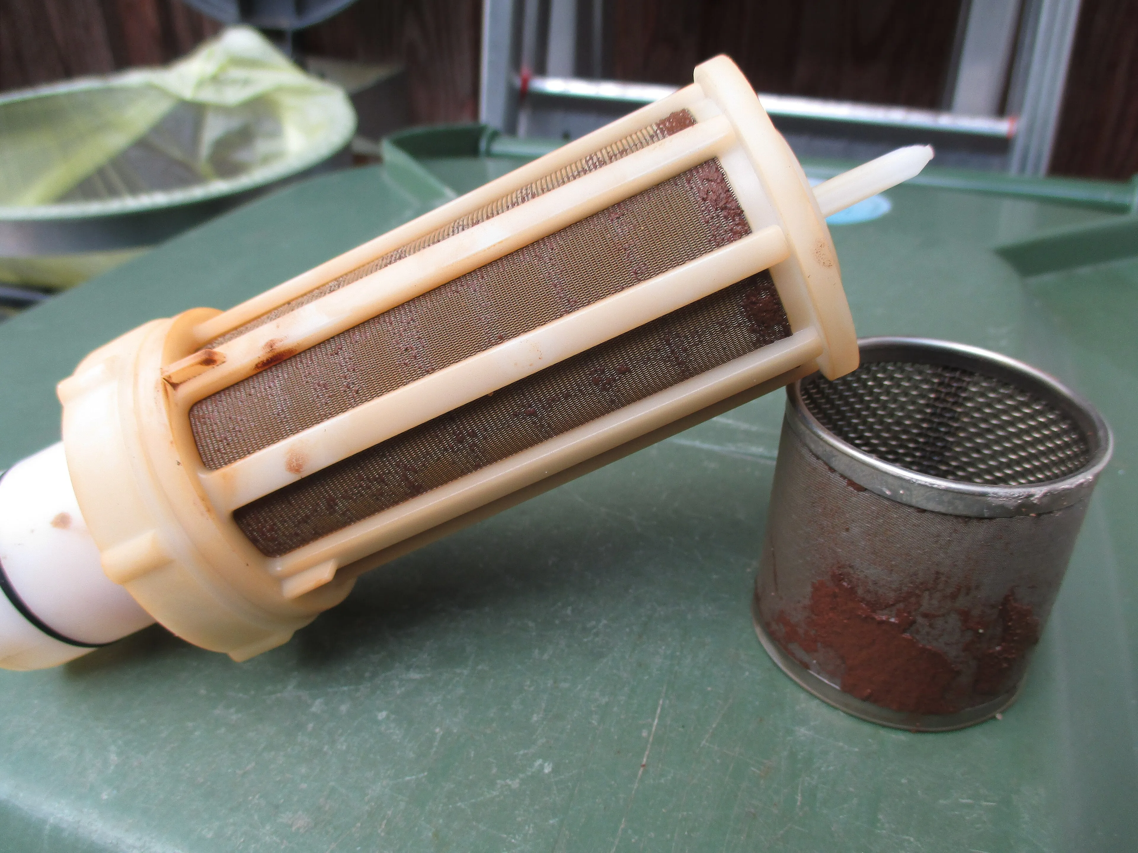 A used water filter cartridge with rust-coloured sediment next to its metal mesh strainer