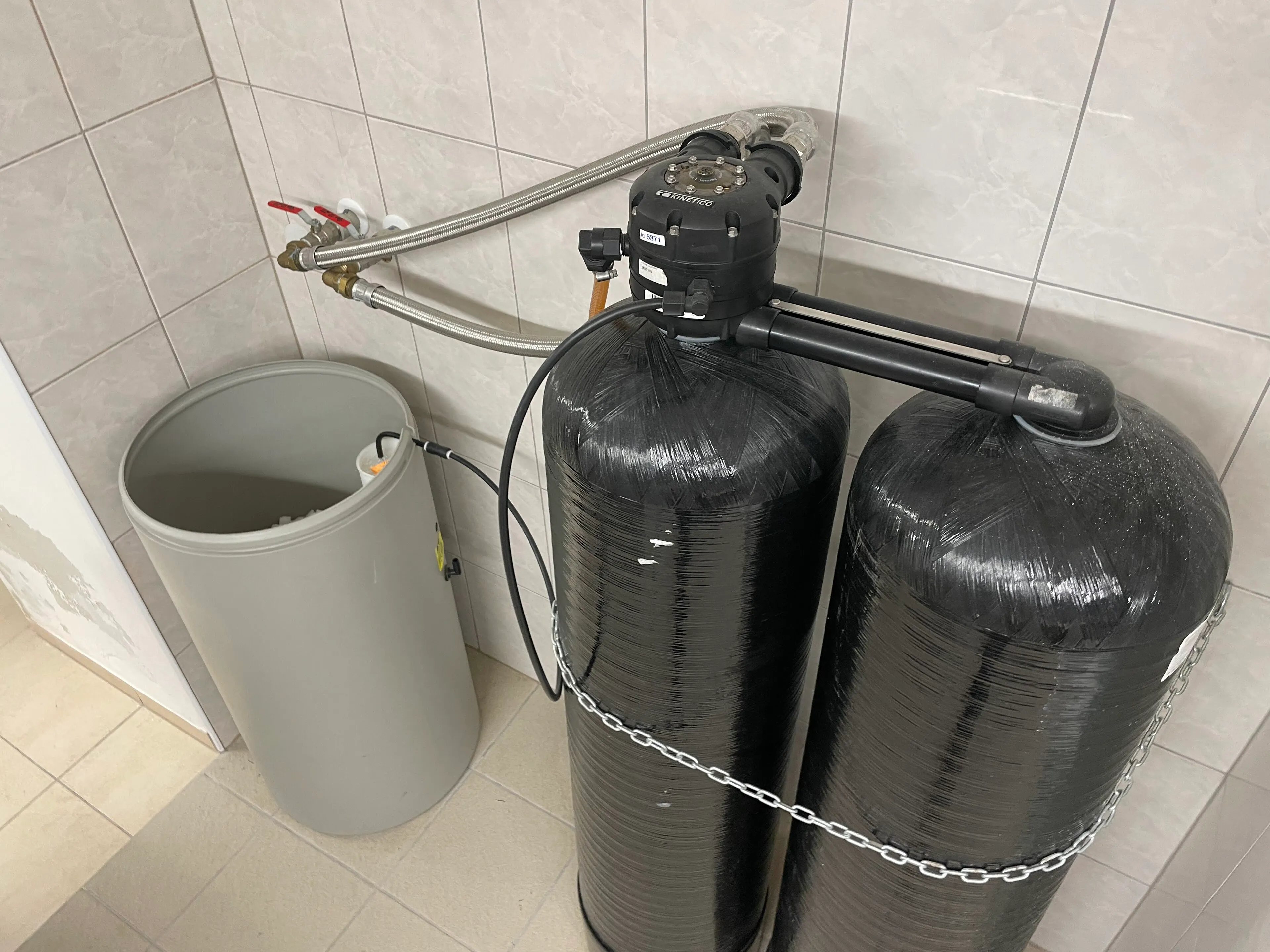 Two black pressure vessels and a brine tank of a water softener installed against a tiled wall